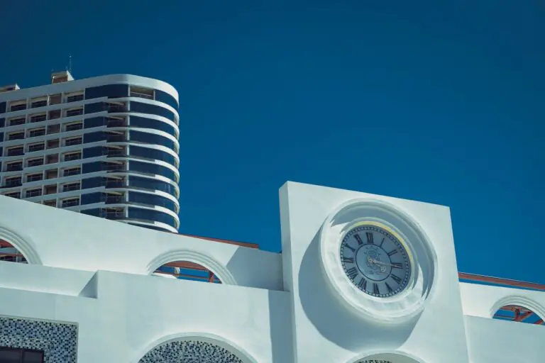 A white building with a large clock face, with a curved high-rise tower against a blue sky