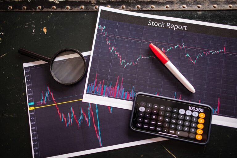 Printed stock charts on a dark table with a magnifying glass, a red marker and a phone calculator
